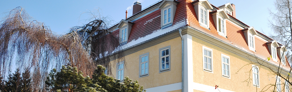 Blick auf einen Teil das Forstamtsgebäudes: gelbe Fassade, kleine Fenster mit Fensterkreuzen und rotem Dach mit Dachfenstern. Davor stehen Bäume