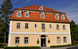 Historisches Forstamtsgebäude mit beiger Fassade und rotem Ziegeldach mit Dachgauben vor strahlend blauem Himmel.