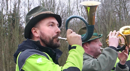 Junger Jagdhornbläser mit Bart, Försterhut und Dienstjacke von ThüringenForst. Im Hintergrund Herbstwald ohne Blätter