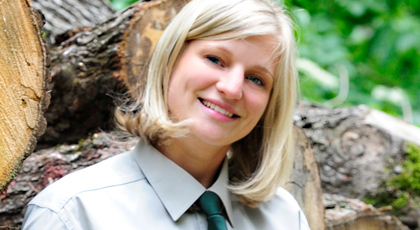 Junge Frau mit forstlicher Dienstkleidung lehnt an einem Holzstapel und lächelt freundlich