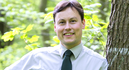 Junger Mann in forstlicher Dienstkleidung, der sich an einen Baum lehnt und freundlich lächelt.