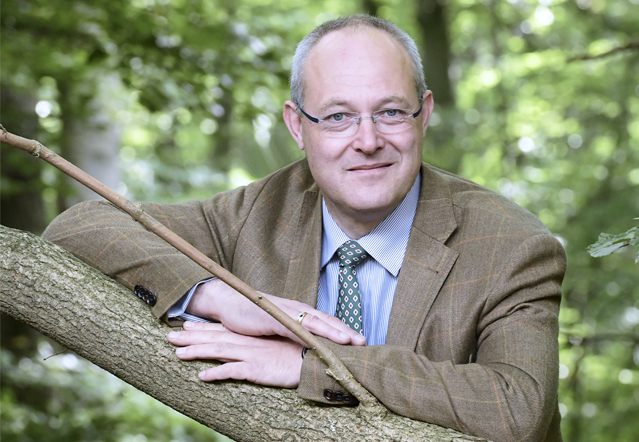 Herr mittleren Alters im Wald. Er lächelt in die Kamera, stützt sich auf einen Ast. der Herr trägt eine braune Blazer-Jacke.