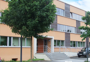 Blick auf ein modernes Bürogebäude im Flachbaustil mit teilweiser Holzverträfelung an der Fassade. Davor stehen kleinere Bäume und es parkt ein Auto
