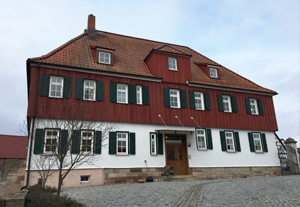 Forsthaus mit weinroter Holzvertäfelung auf halber Haushöhe. Das Dach ist ebenfalls weinrot, die untere Gebäude-Etage trägt hellen Putz