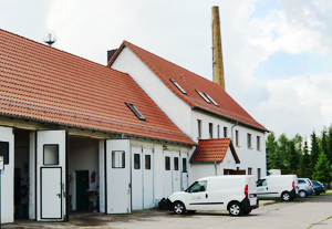 Gebäude mit rotem Spitzdach, davor parken Autos und ein Tor zur Einfahrt für große Fahrzeuge steht offen