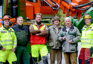 Gruppe von 6 Männern in neonfahrenber Arbeitskleidung und forstlichen Uniformen steht freundlich schauend vor einem LKW-ähnlichem Holzerntefahrzeug