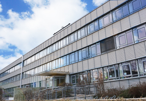 Blick auf ein modernes Gebäude im Flachbaustil mit großen Fensterfronten, in denen sich der blaue Himmel spielgelt
