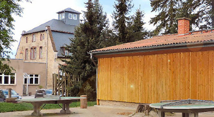 Gebäude im altertülichen Villen-Baustil mit Natursteinfassade, vor ein Hoilz-Flachbau mit Spielplatz