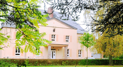Gebäude im historischen Villen-Stil mit lachsfarbener Fassade, eingerahmt von Laub- und Nadelbäumen