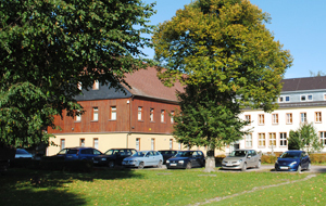 Blick auf ein Eckgebäude, teilweise mit Holzvertäfelung, davor parkende Autos und grüne Bäume