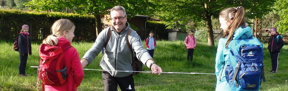 Ein Förster steht mit einem Zollstock bei im Kreis stehenden Kindern und hält den Zollstock vor den Kindern.