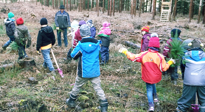 Ein Forster ist mit einer Gruppe von Kindern im Wald unterwegs. Die Kinder tragen bunte Jacken und Regenhosen.
