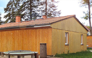 Flachbau mit Holzfassade, Tischtennisplatte, dahinter zahlreiche Kiefernbäume