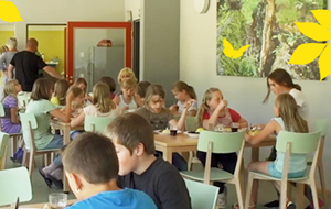 Viele Kinder sitzen in einem mordern eingerichten Speisesaal. Tische und Stühle sind zartgrün und holzfarben. An der Wand hängen Waldbilder.