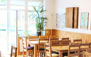 Einladend aussehender Raum in Holzoptik mit Holztischen und Stühlen. An der Wand: halbhohe Holzvertäfelung. Der Raunm öffnet sich zum Hof durch eine große Glastür. 