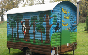 Mit bunten Waldmotiven bemalter Bauwagen aus Holz steht auf einer Wiese.
