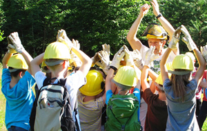 Gruppe von Kindern mit Lehrerin: alle tragen einen gelben "Baurbeiterhelm" und halten ihre Hände in die Luft, als wären Sie Bäume, die sich im Wind wiegen